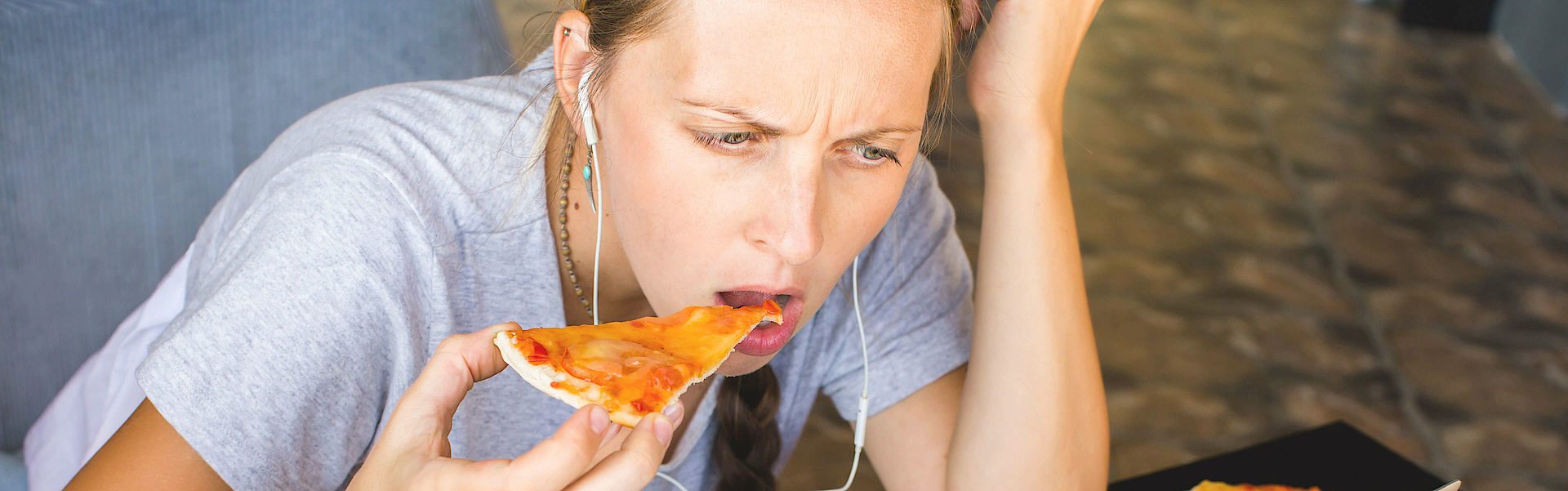 Eine Frau sitzt vor einem Laptop. Sie schaut auf den Bildschirm und isst ein Stück Pizza. Um sie herum liegt einiges an Fast Food und ungesundem Essen.