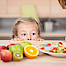 Kleines Kind etwas größer als der Küchentisch kann gerade so den Obstteller auf diesem betrachten. Am rechten Rand des Bildes sieht man Umrisse einer Frau.
