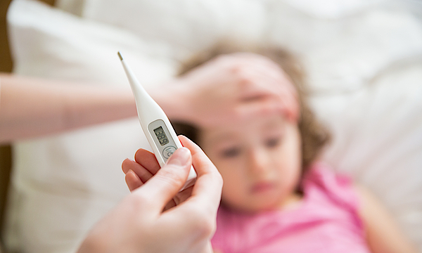 Ein kranken Kind liegt im Bett. Es hat hohes Fieber. Die Mutter hält ein digitales Fiebertermometer. Das ist im Closeup zu sehen.