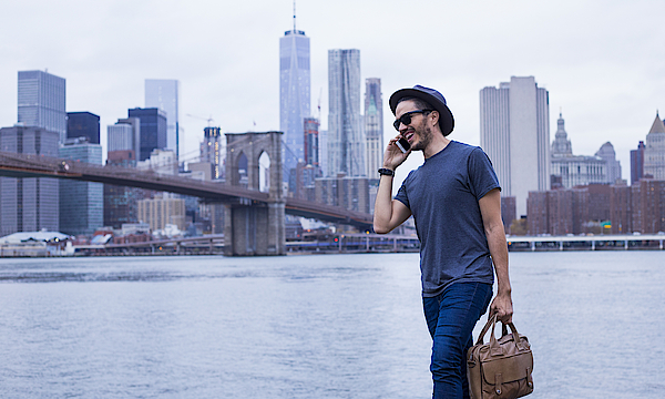 Ein Mann vor der Skyline von New York. Er trägt eine Laptop-Tasche und telefoniert.
