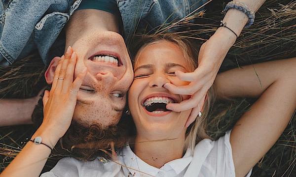 Ein Junge und ein Mädchen liegen auf einer Wiese, Kopf an Kopf. Sie sind Teeanger. Beide lachen. Der Junge schaut das Mädchen an. Sie hat die Augen geschlossen. Sie hat eine feste Zahnspange.