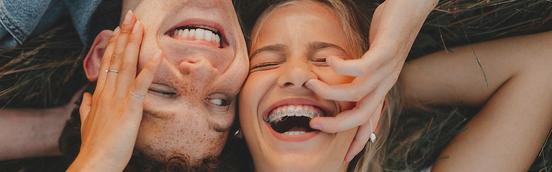 Ein Junge und ein Mädchen liegen auf einer Wiese, Kopf an Kopf. Sie sind Teeanger. Beide lachen. Der Junge schaut das Mädchen an. Sie hat die Augen geschlossen. Sie hat eine feste Zahnspange.