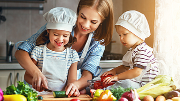 Die Haushaltshilfe der Familie bereitet mit den Kindern das Essen vor. Das größere Kind hilft mit und das kleine schaut zu.