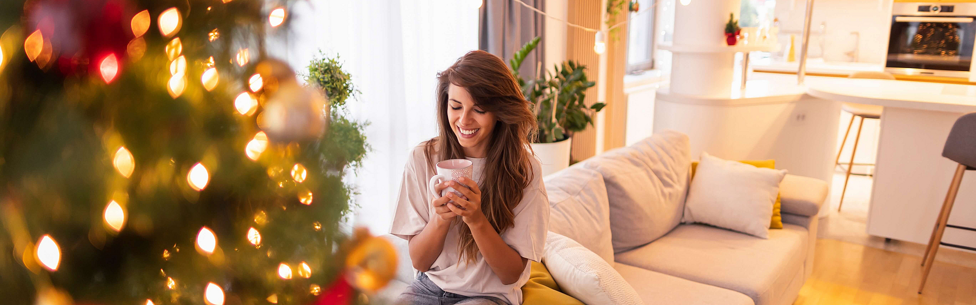 Zu sehen ist eine Frau im Wohnzimmer mit einer Tasse vor dem Weihnachtsbaum.