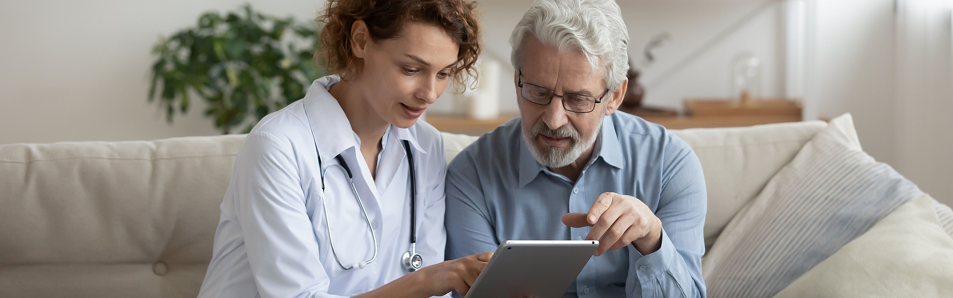 Eine Ärztin und ihr alterer Patient sitzen auf einem Sofa. Die Arztin hält ein Tablett. Sie erklärt dem Patienten die Ergebnisse einer Untersuchtung.