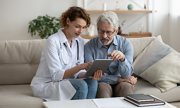 Eine Ärztin und ihr alterer Patient sitzen auf einem Sofa. Die Arztin hält ein Tablett. Sie erklärt dem Patienten die Ergebnisse einer Untersuchtung.