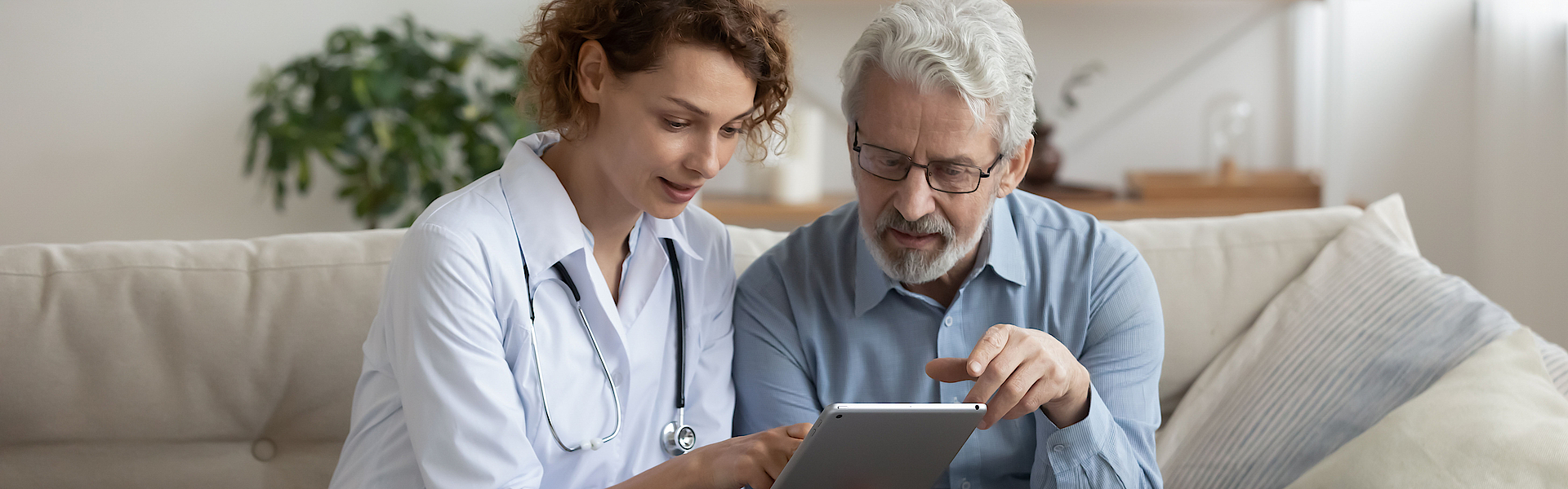 Eine Ärztin und ihr alterer Patient sitzen auf einem Sofa. Die Arztin hält ein Tablett. Sie erklärt dem Patienten die Ergebnisse einer Untersuchtung.