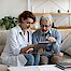 Eine Ärztin und ihr alterer Patient sitzen auf einem Sofa. Die Arztin hält ein Tablett. Sie erklärt dem Patienten die Ergebnisse einer Untersuchtung.