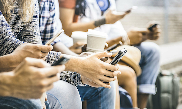 Eine Gruppe junger Menschen sitzt auf einer Mauer. Sie alle haben Smartphones in der Hand und schauen darauf. 