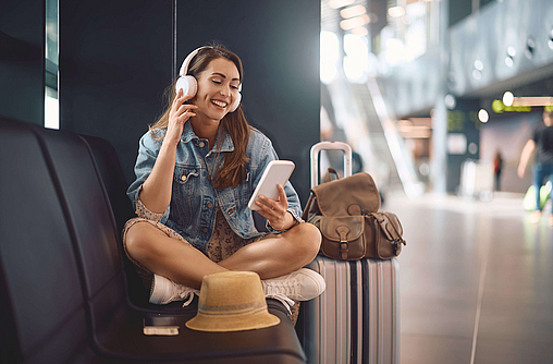 Frau sitzt im Abflugbereich auf dem Weg in den Urlaub. Sie hört Musik über das Smartphone.