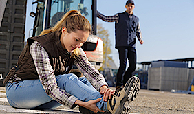 Eine Frau sitzt auf dem Boden und hält sich den Knöchel fest. Im Hintergrund stehen ein Baustellenfahrzeug und ein Arbeiter.