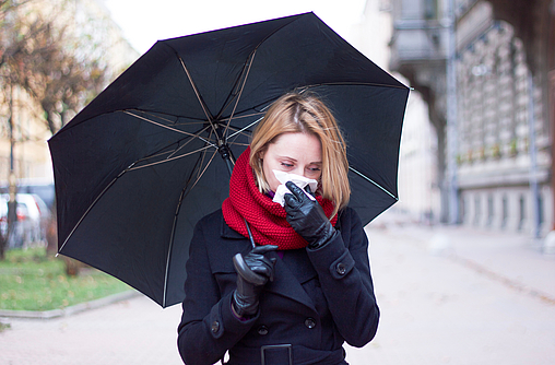 Frau im dunklen Mantel mit Regenschirm und Schnupfen läuft durch eine Straße.