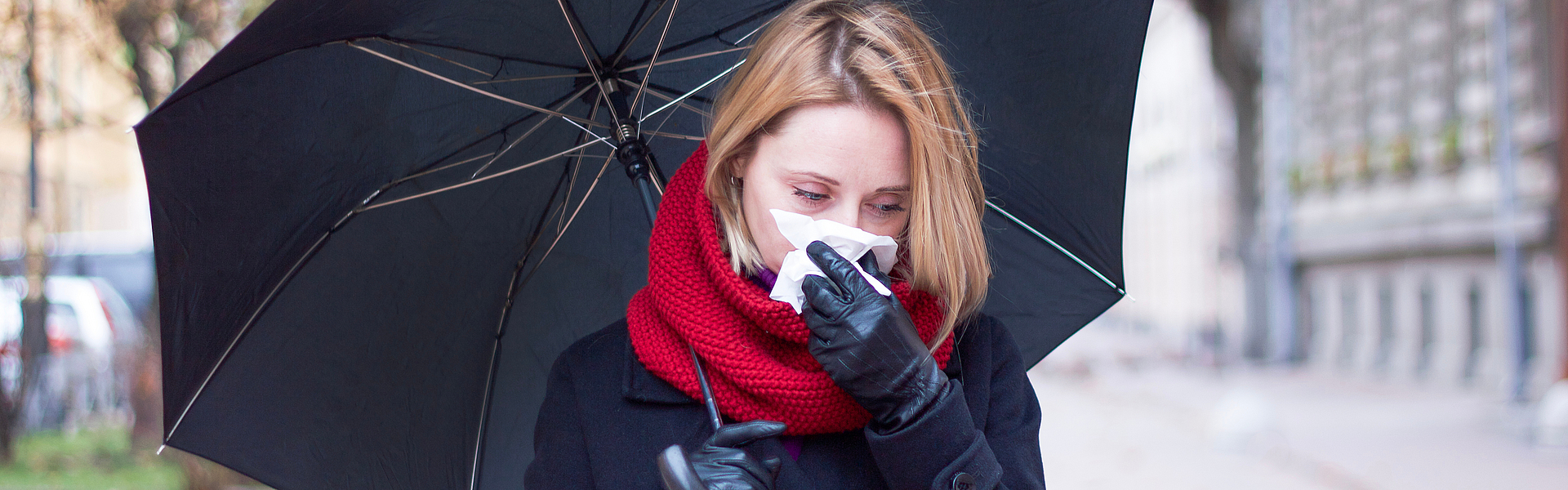 Frau im dunklen Mantel mit Regenschirm und Schnupfen läuft durch eine Straße.