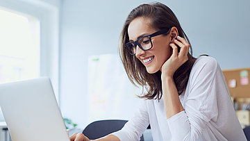 Lächelnde junge Frau sitzt am Schreibtisch und arbeitet am Laptop.