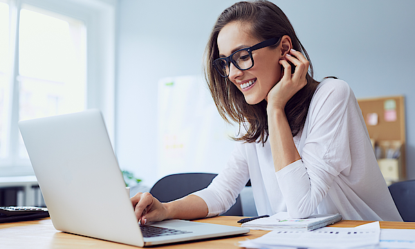 Lächelnde junge Frau sitzt am Schreibtisch und arbeitet am Laptop.