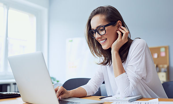 Lächelnde junge Frau sitzt am Schreibtisch und arbeitet am Laptop.