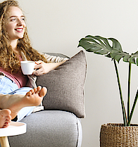 Lächende Frau sitzt auf dem Sofa und hat die Beine auf den Tisch gelegt. Sie hält eine Kaffetasse in der linken Hand.