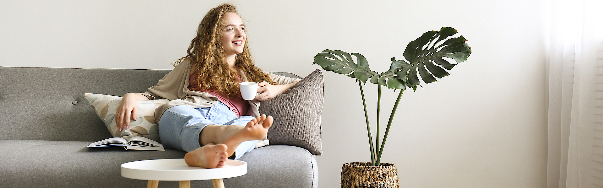 Lächende Frau sitzt auf dem Sofa und hat die Beine auf den Tisch gelegt. Sie hält eine Kaffetasse in der linken Hand.