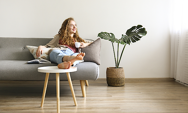 Lächende Frau sitzt auf dem Sofa und hat die Beine auf den Tisch gelegt. Sie hält eine Kaffetasse in der linken Hand.