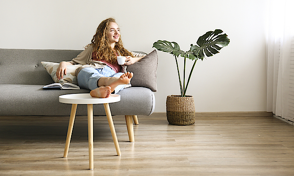 Lächende Frau sitzt auf dem Sofa und hat die Beine auf den Tisch gelegt. Sie hält eine Kaffetasse in der linken Hand.