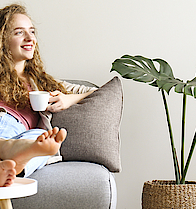 Lächende Frau sitzt auf dem Sofa und hat die Beine auf den Tisch gelegt. Sie hält eine Kaffetasse in der linken Hand.