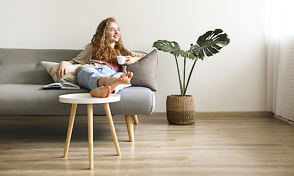 Lächende Frau sitzt auf dem Sofa und hat die Beine auf den Tisch gelegt. Sie hält eine Kaffetasse in der linken Hand.