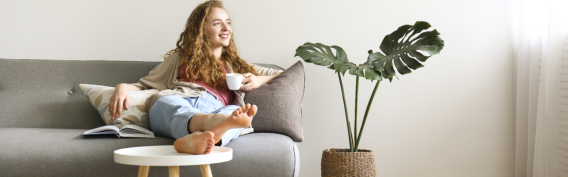 Lächende Frau sitzt auf dem Sofa und hat die Beine auf den Tisch gelegt. Sie hält eine Kaffetasse in der linken Hand.