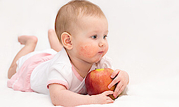 Ein Baby liegt auf dem Bauch und hält einen roten Apfel fest. Die Wangen des Kindes sind aufgrund einer Allergie gerötet.