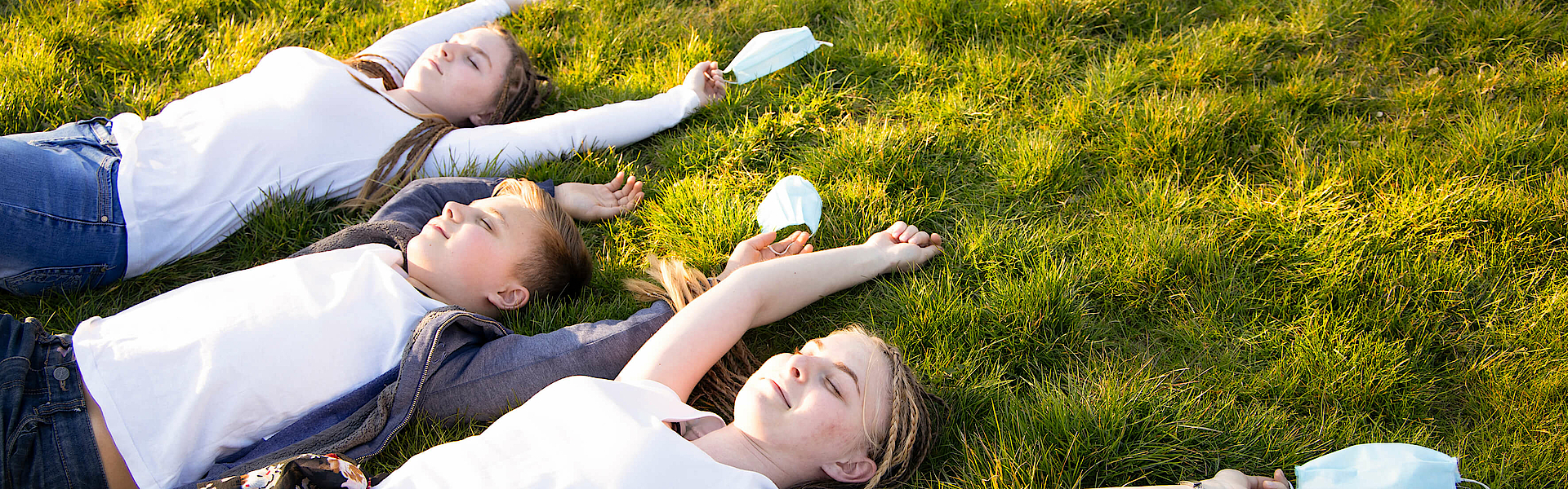 Zwei Kinder und eine Jugendliche liegen auf einer grünen Wiese. Sie genießen die Luft und atmen durch. Ihre Arme sind von ihnen gestreckt. In ihren rechten Händen halten sie medizinische Masken. Sie freuen sich, dass sie die Masken nicht tragen müssen. Alle drei lachen und sind glücklich.