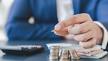 Mann hält Euro-Münze in der Hand, auf dem Tisch liegt ein Taschenrechner und Geld.