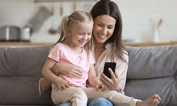 Eine Mutter sitzt mit ihrer kleinen Tochter auf dem Sofa. Die Mutter hat ein Smartphone in der Hand.