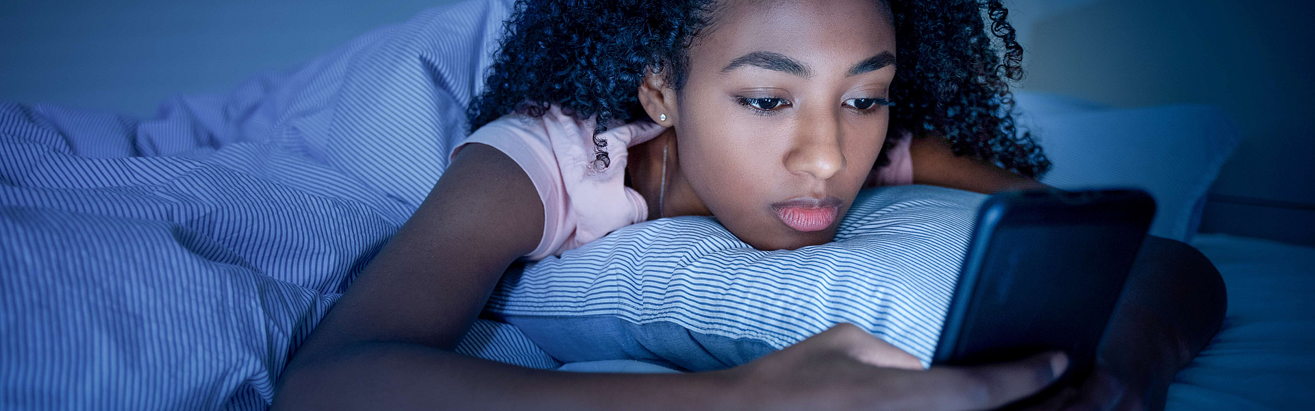 Eine junge Frau liegt im Dunkeln auf dem Bett und schaut auf ihr beleuchtetes Smartphone