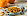 Kürbiskerne liegen auf einem Teller. Der Teller steht auf einem Tuch. Neben dem Teller liegen ein Löffel und weitere Kürbiskerne. Im Hintergrund befinden sich zwei Kürbisse als Dekoration. 