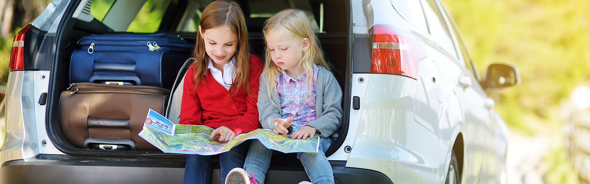 Die gepackten Koffer sind im Auto, zwei Kinder sitzen im Kofferraum und schauen auf eine Wegbeschreibungskarte.