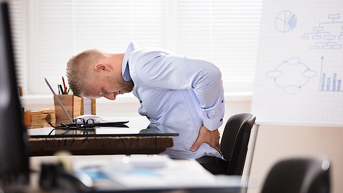 Ein Mann sitzt im Büro an seinem Schreibtisch. Er ist vorn übergebeugt und hält sich den Rücken. Er hat Schmerzen. Sein Gesicht ist schmerzverzerrt.