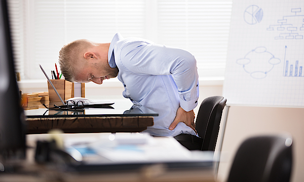 Ein Mann sitzt im Büro an seinem Schreibtisch. Er ist vorn übergebeugt und hält sich den Rücken. Er hat Schmerzen. Sein Gesicht ist schmerzverzerrt.