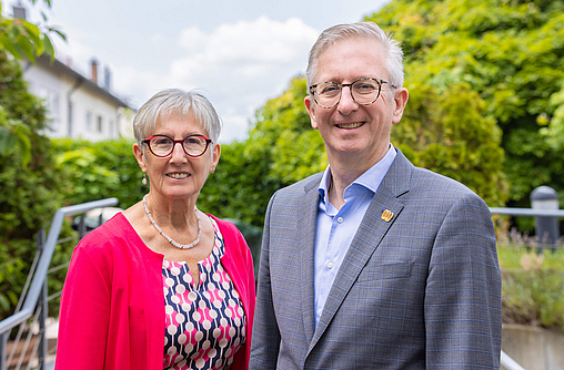 Die Verwaltungsratsvorsitzenden der mhplus, Christine Wagner und Christian Ottenbacher, stehen nebeneinander auf einer Treppe mit Bäumen im Hintergrund. 
