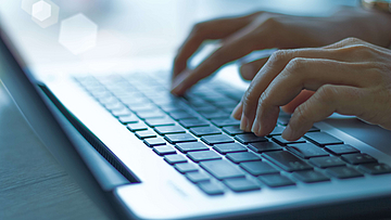 Auf dem Tisch liegt ein offener Laptop, zwei Hände betätigen die Tastatur.