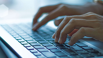 Auf dem Tisch liegt ein offener Laptop, zwei Hände betätigen die Tastatur.