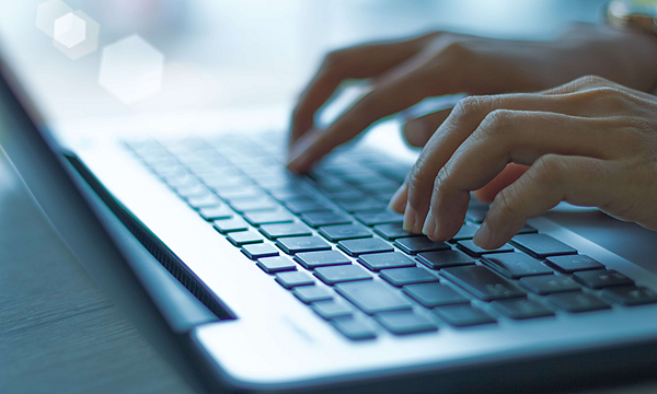 Auf dem Tisch liegt ein offener Laptop, zwei Hände betätigen die Tastatur.
