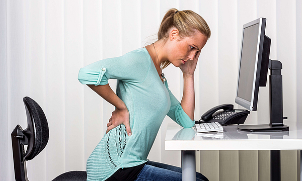 Eine Frau sitzt an einem Schreibtisch und hat Rückenschmerzen.