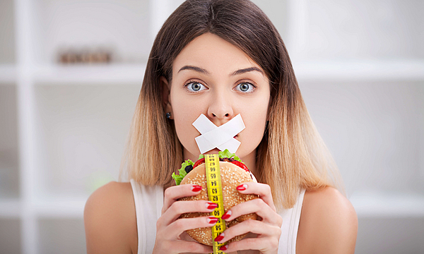 Zu sehen ist eine Frau, der ihr Mund zugeklebt wurde und einen Burger mit beiden Händen hält.