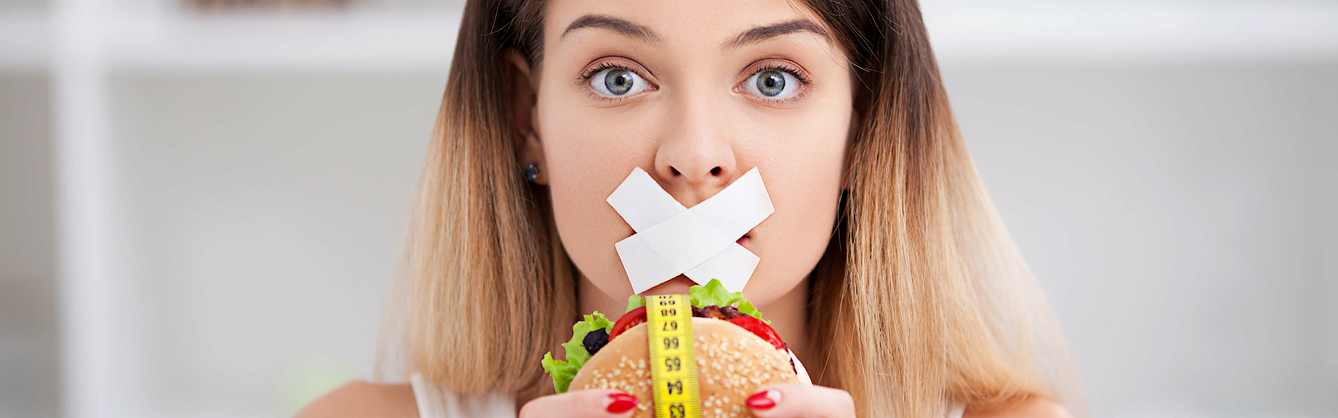 Zu sehen ist eine Frau, der ihr Mund zugeklebt wurde und einen Burger mit beiden Händen hält.