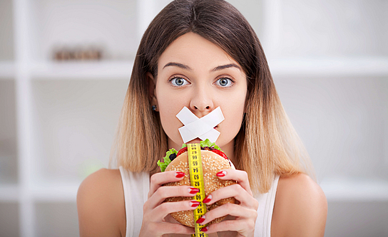 Zu sehen ist eine Frau, der ihr Mund zugeklebt wurde und einen Burger mit beiden Händen hält.