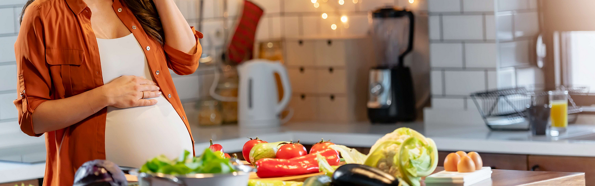 Schwangere Frau steht in der Küche. Auf der Arbeitsfläche liegt Salat mit vielen Vitaminen. Sie isst lachend einen Paprika. 