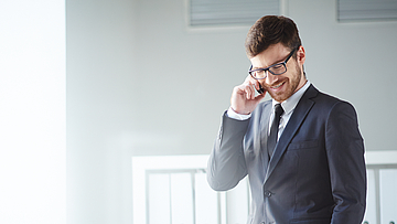 Ein Mann steht im Büro und telefoniert mit dem Handy. Er hat eine Hand in der Hosentasche.