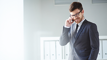 Ein Mann steht im Büro und telefoniert mit dem Handy. Er hat eine Hand in der Hosentasche.