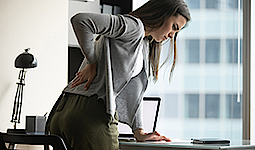Eine junge Frau mit grüner Hose und grauer Strickjacke stützt sich mit einer Hand auf ihrem Schreibtisch ab. Die andere Hand ist auf ihrem Rücken, weil sie Rückenschmerzen hat.