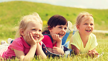 Drei kleine Kinder liegen auf einer Yogamatte auf einer grünen Wiese. Die Sonne scheint. Die Kinder sind glücklich und lachen. 