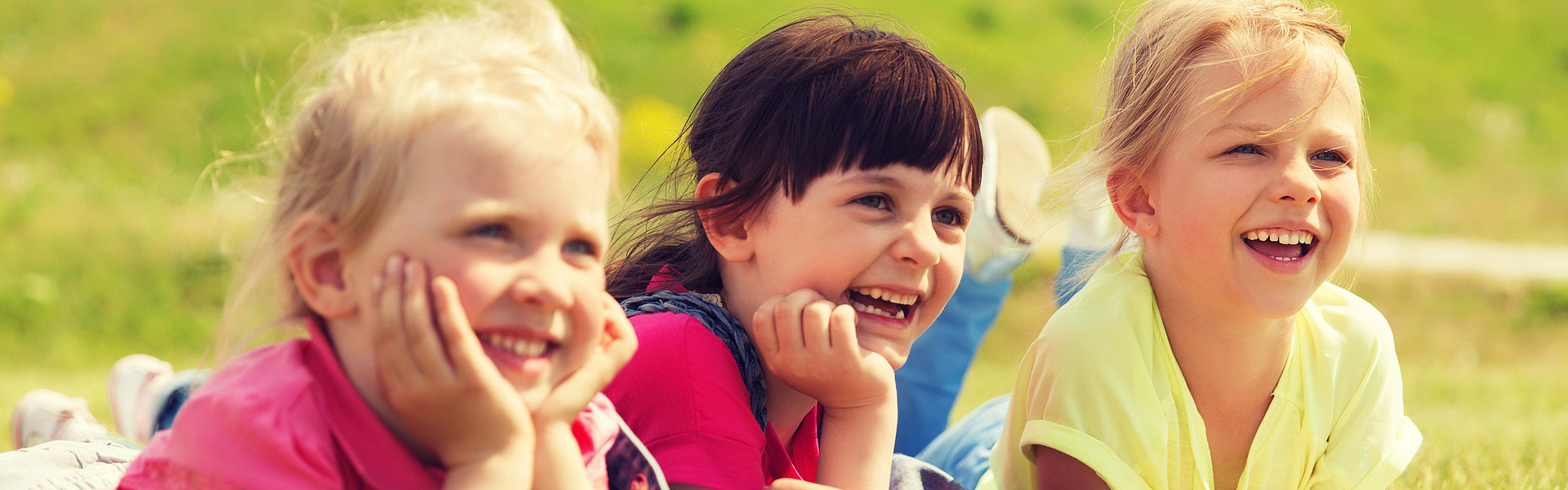 Drei kleine Kinder liegen auf einer Yogamatte auf einer grünen Wiese. Die Sonne scheint. Die Kinder sind glücklich und lachen. 